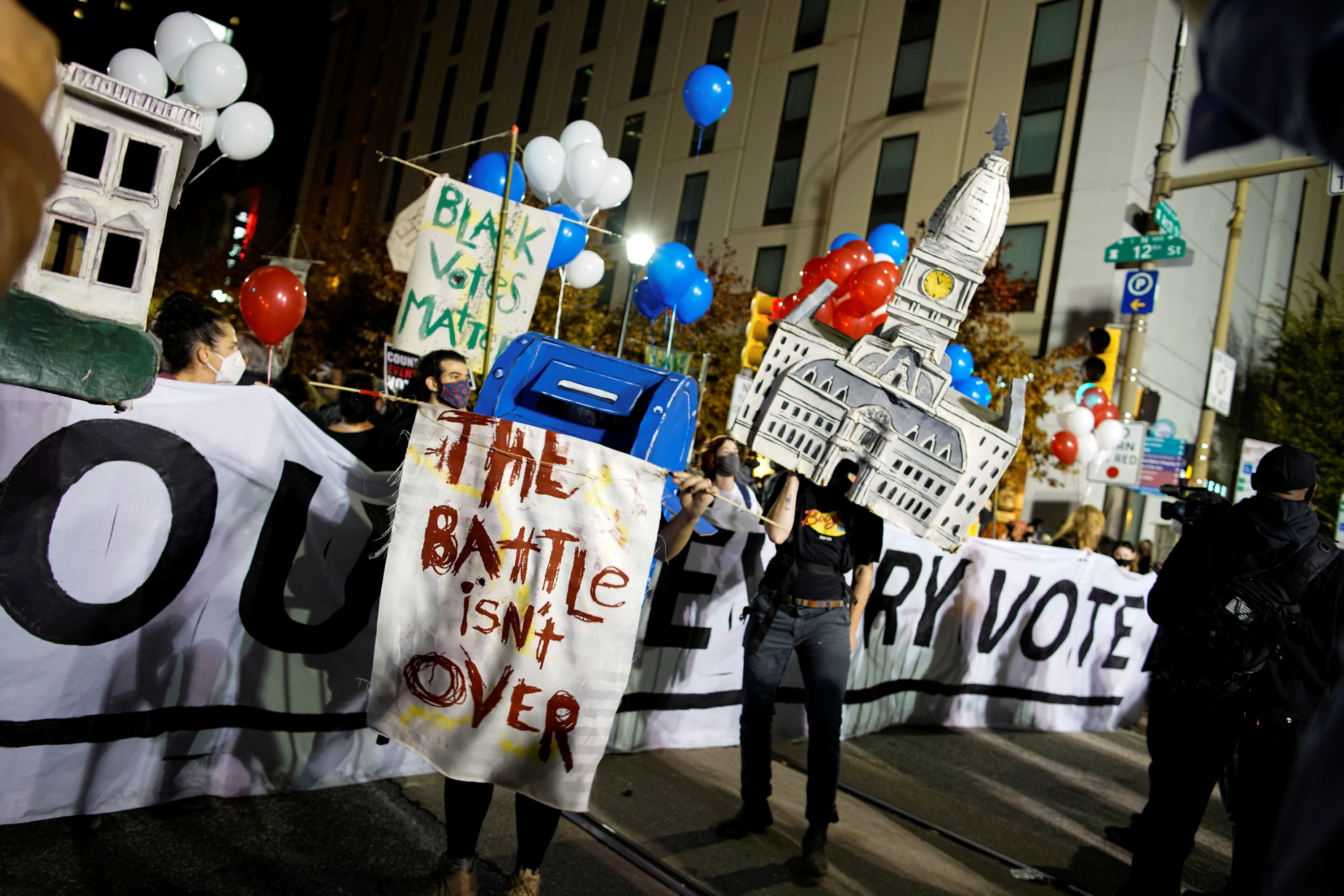 Person wearing a mailbox costume holds sign saying, &quot;The Battle isn&#x27;t Over&quot;