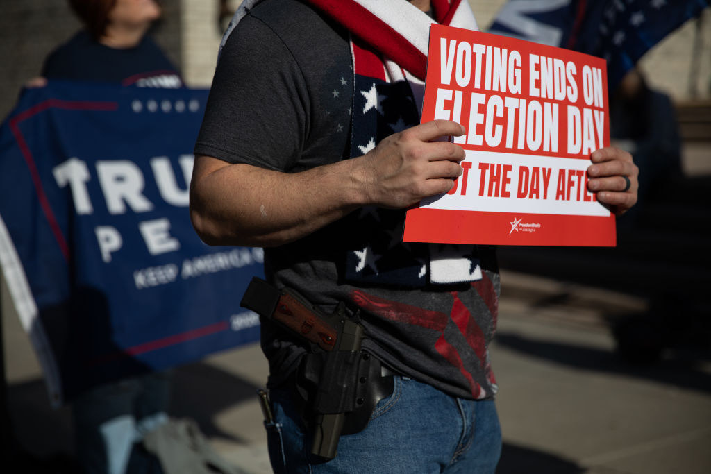 Man with a gun in a holster holds sign saying &quot;voting ends on Election Day, Not the day after&quot;
