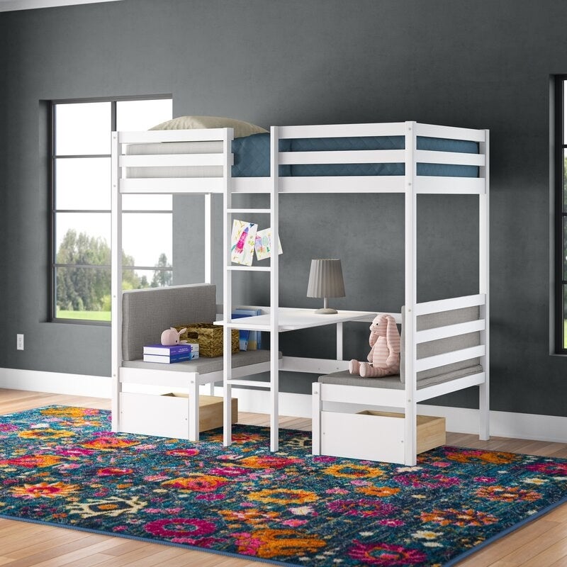 A white loft bed with a bed at the top bunk and a table and bench set below on top of a floral rug