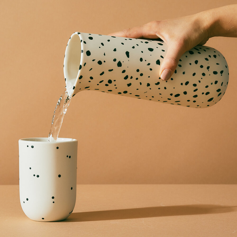 The white pitcher with black spots on it pouring water into a cup