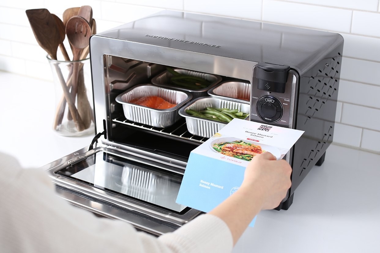 A person&#x27;s hand reads operating instructions for a smart oven that sits atop a white counter next to a clear utensil crock with wooden spoons