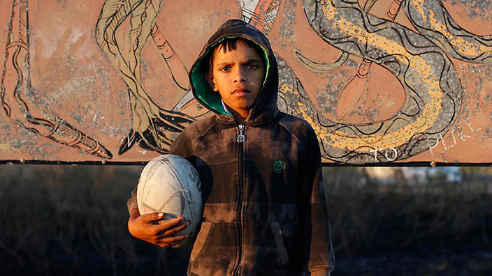 A young boy standing in front of a mural, holding a rugby ball