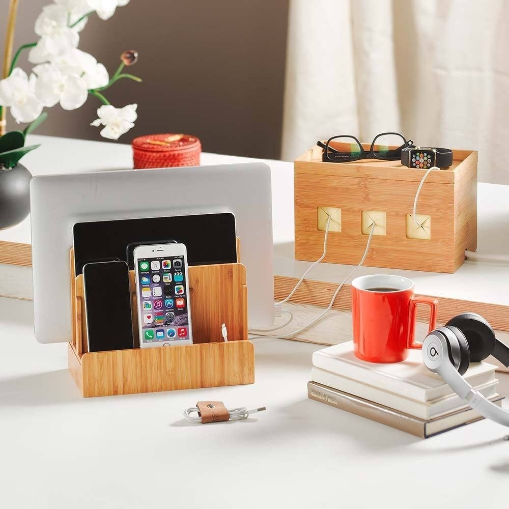 Wooden device charging station on a multilevel surface alongside wires, books, flowers, headphones, a container, and a mug of coffee