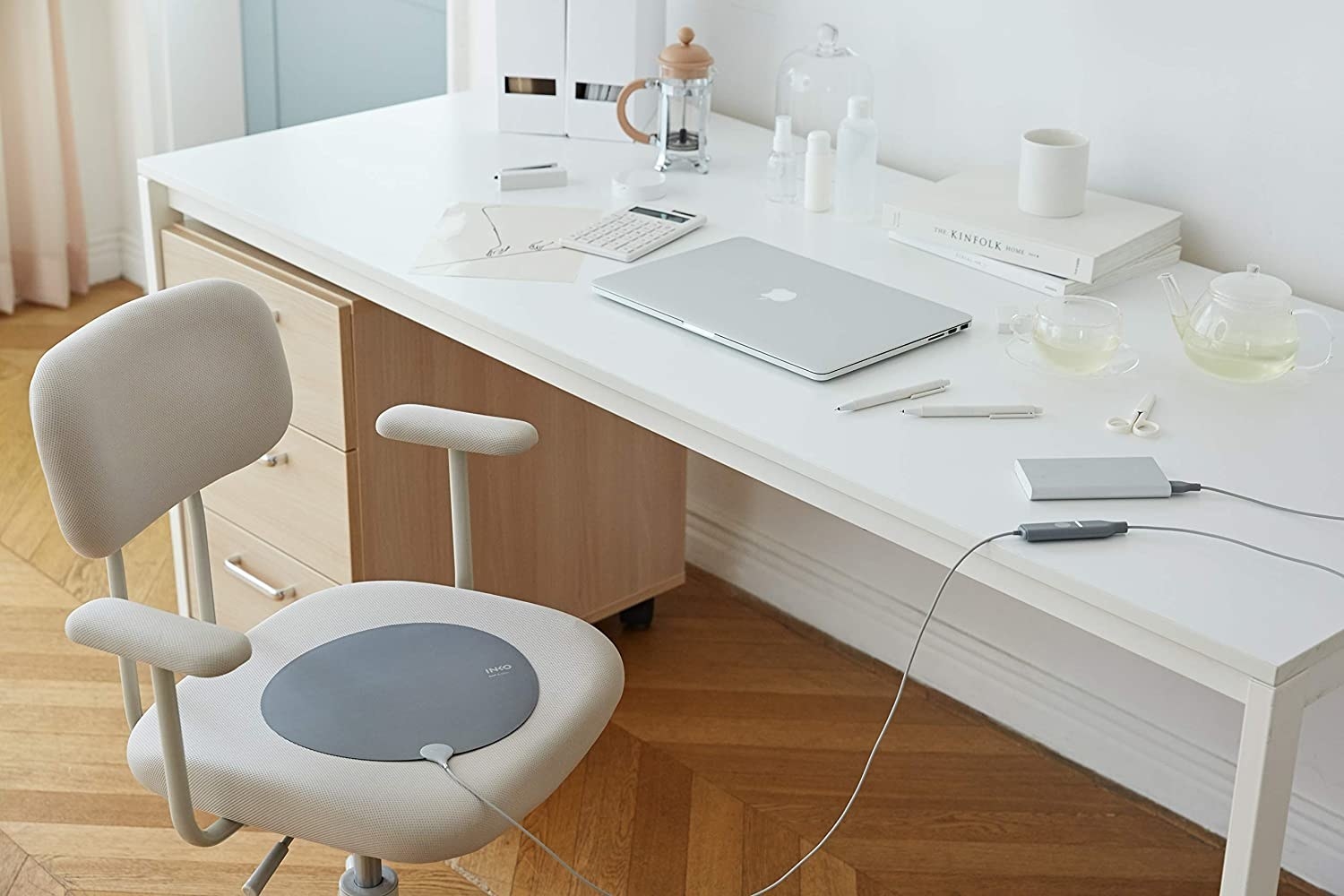 Home office setup with a white, light brown, and cream aesthetic, including a laptop, books, scissors, calculator, French press, gray heating pad, and more