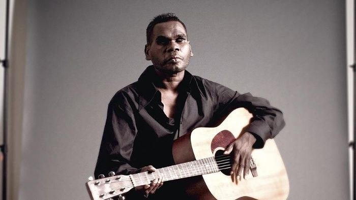 Gurrumul sitting down on a stool and holding his guitar