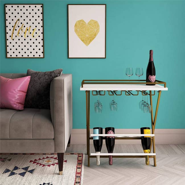 A gold bar cart with a white top and gold hinges at the bottom to hold wine glasses 