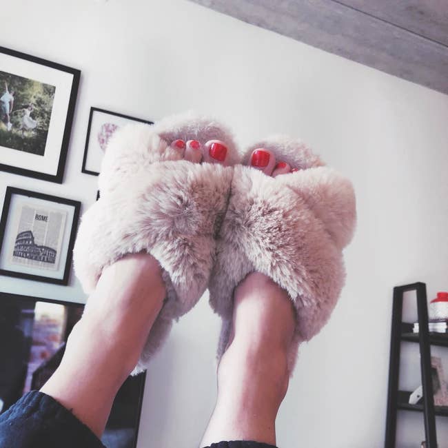 Reviewer pic of their feet in the air with the fuzzy slippers on with a criss-cross over the top of the foot and an open-toe style in light pink