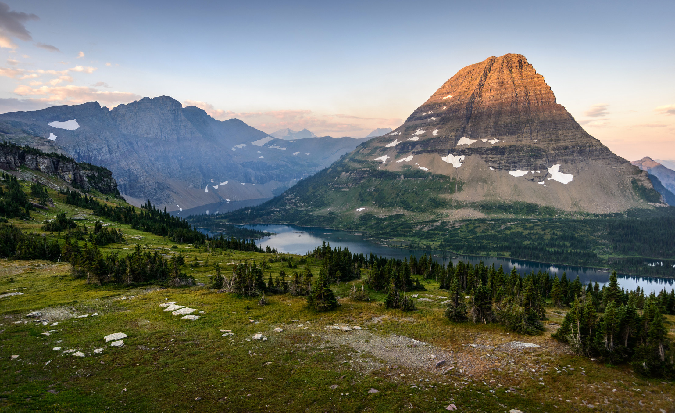 A wide shot of a cliff dotted with trees overlooking a lake and a tall mountain in Glacier National Park