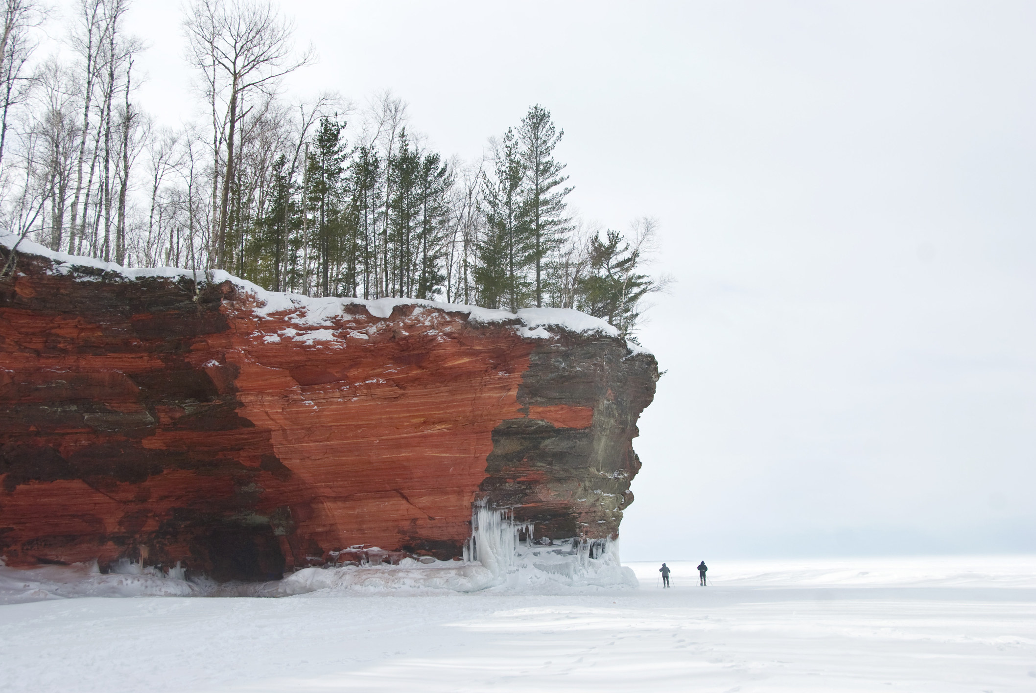 Red, sandstone cliffs jut out over a frozen lake where two small figures walk