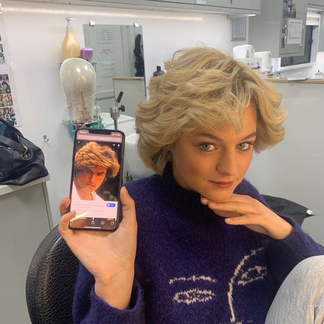 Emma Corrin sitting in a dressing room trailer; she has her hand tucked under her chin as she points her phone at the camera; her phone has an image of a blonde-haired man with a shaggy hairstyle