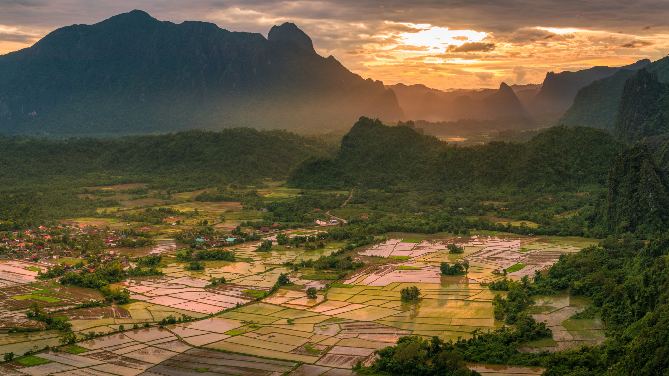 A view over rice paddy fields with big rocky mountains behind them at sunset