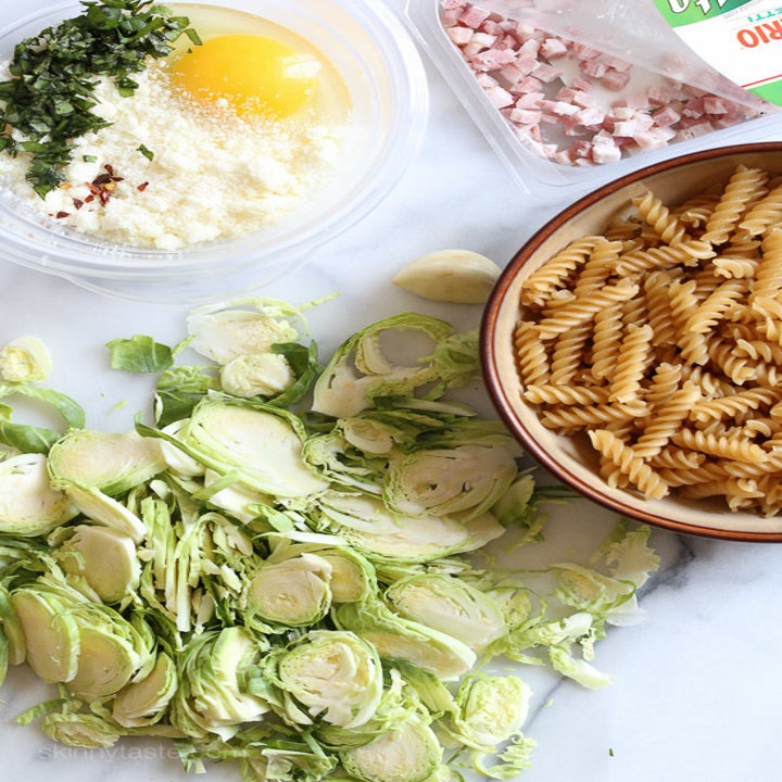 Brussels sprouts, pasta, pancetta, cheese, and cracked egg on a countertop.
