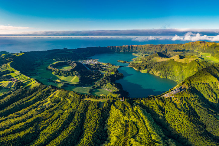 Aerial shot of a lush green island shaped like a volcano, with bright blue water in the middle