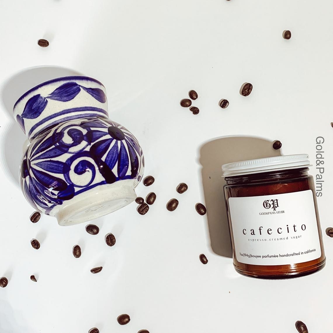 A cafecito candle laying next to spilled coffee beans and a blue and white majolica cup