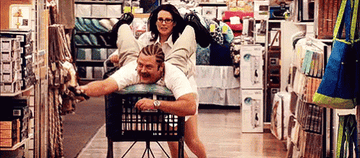 Tammy wheeling Ron down the aisles of a store in a shopping cart as he scans items for their registry