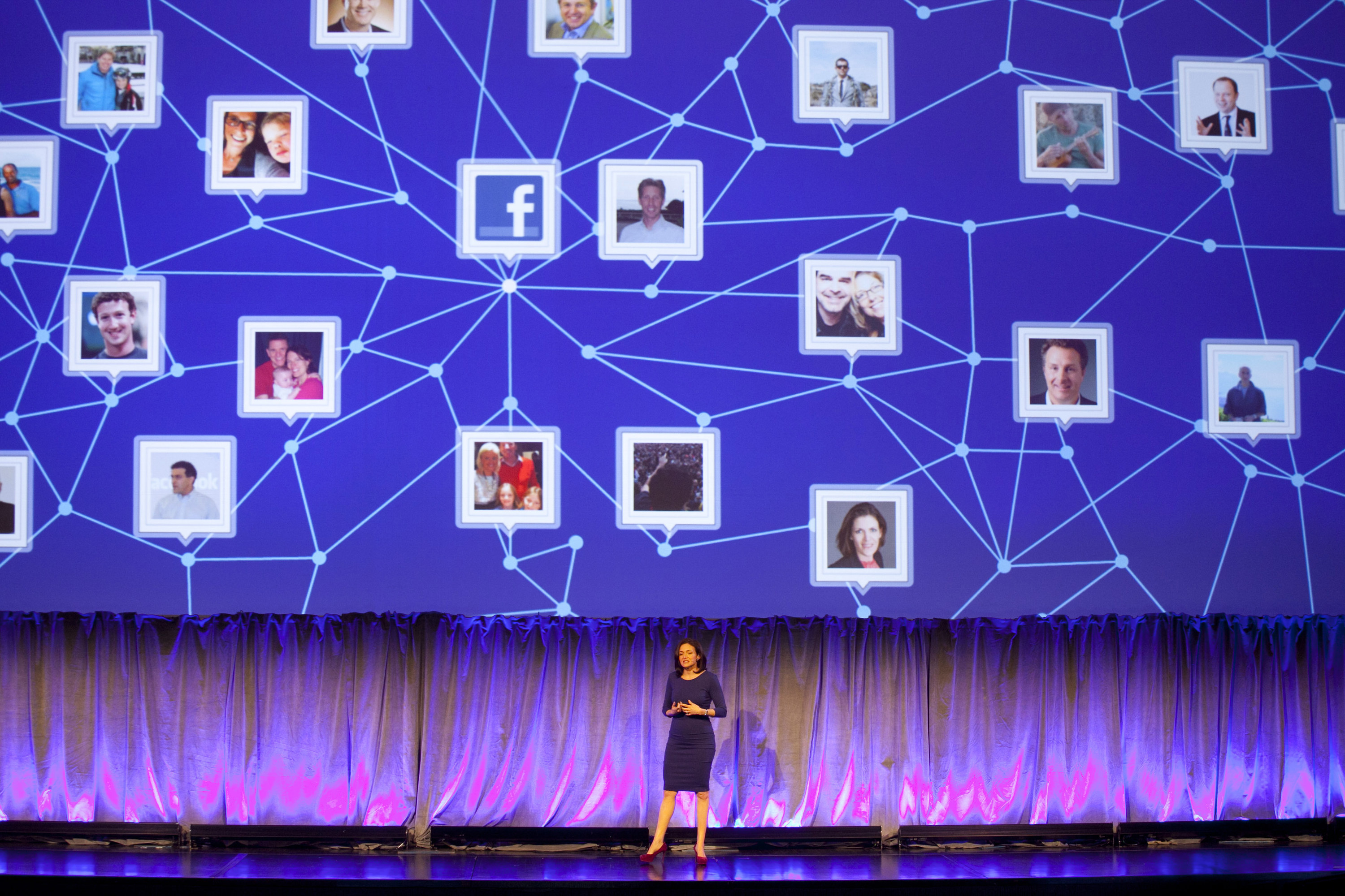 A woman speaks onstage in front of a large diagram above her that shows images of people connected to one another by lines, resembling a vast social network