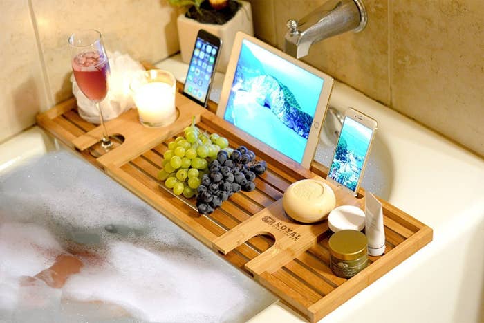 the bath tray displayed over a bath-tub with wine, grapes, a candle, and technology devices on it