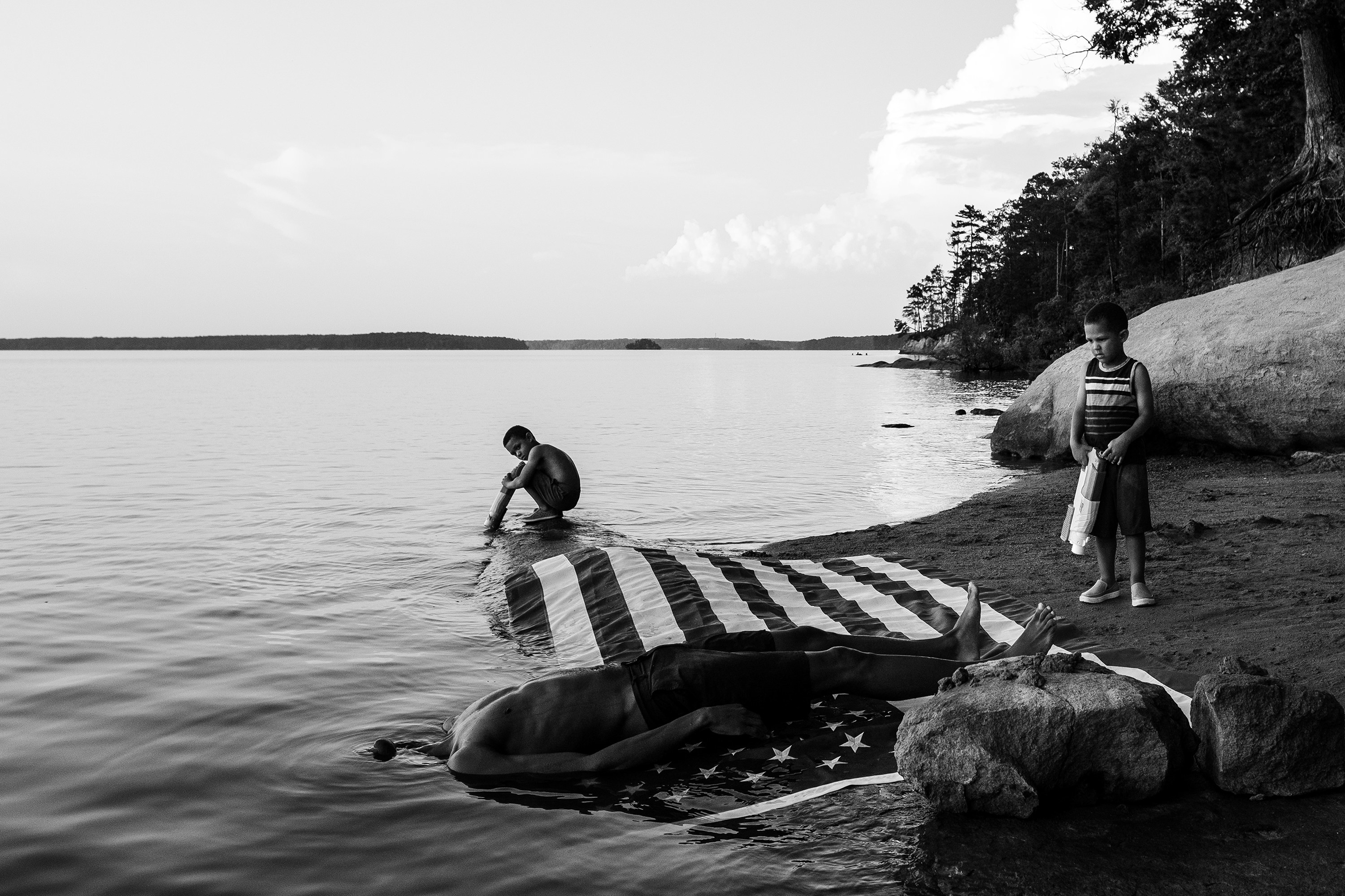 A man lying on a flag with his head underwater as two boys watch him