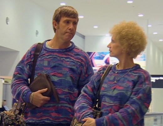 Kath and Kel stand in matching sweaters in the airport, looking at each other