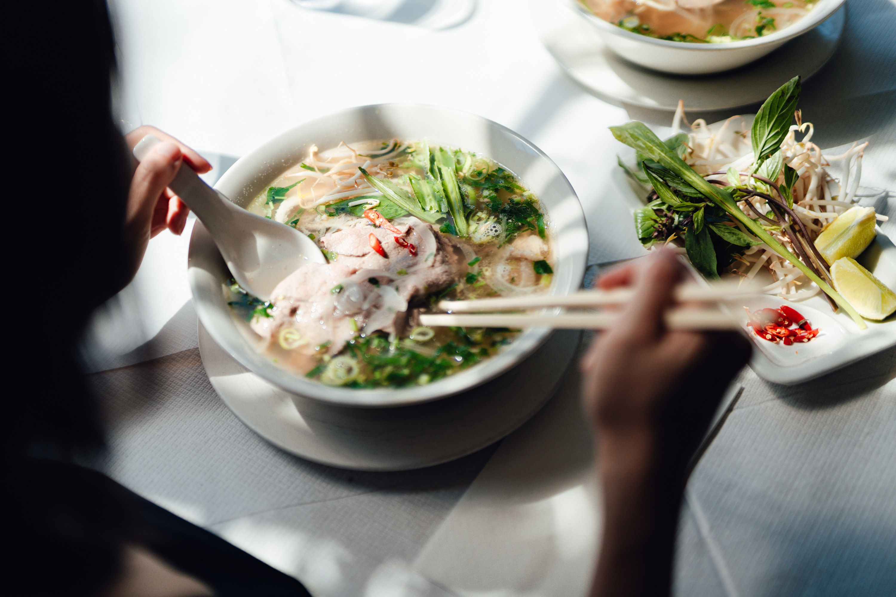 Over-the-shoulder view of young woman eating traditional Vietnamese pho soup with chopsticks.