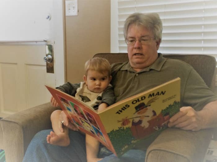 George Finefrock sits in an armchair with his infant grandson on his lap, holding open a large picture book in front of them titled &quot;This Old Man&quot;