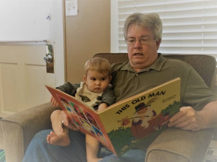 George Finefrock sits in an armchair with his infant grandson on his lap, holding open a large picture book in front of them titled "This Old Man"