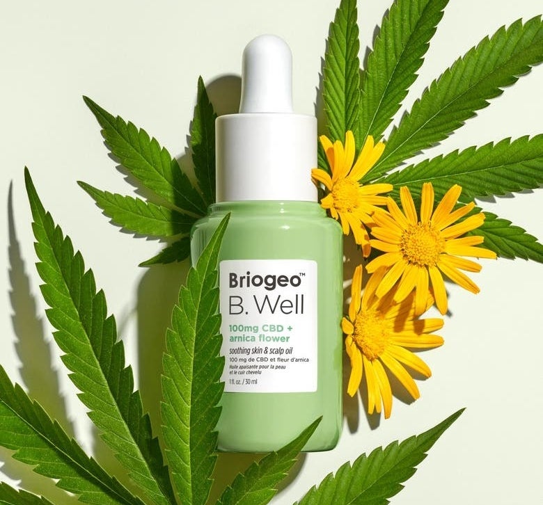 A green bottle of Briogeo B.Well skin and scalp oil surrounded by marijuana leaves and yellow arnica flowers 