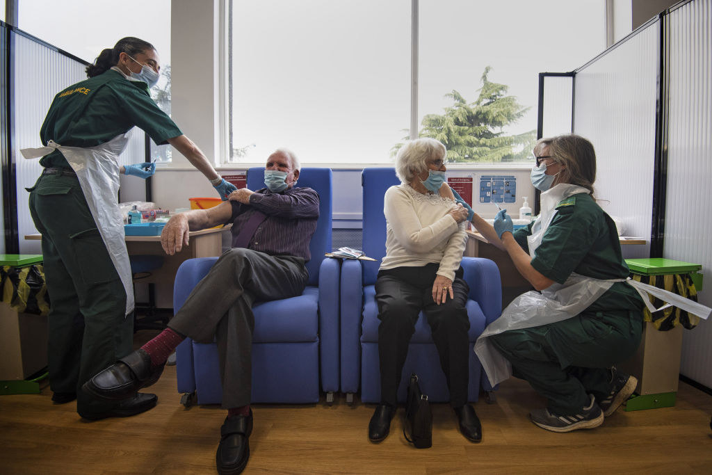 An older man and an older woman sit in cushioned seats in face masks as they receive COVID-19 vaccines from two nurses in green uniforms