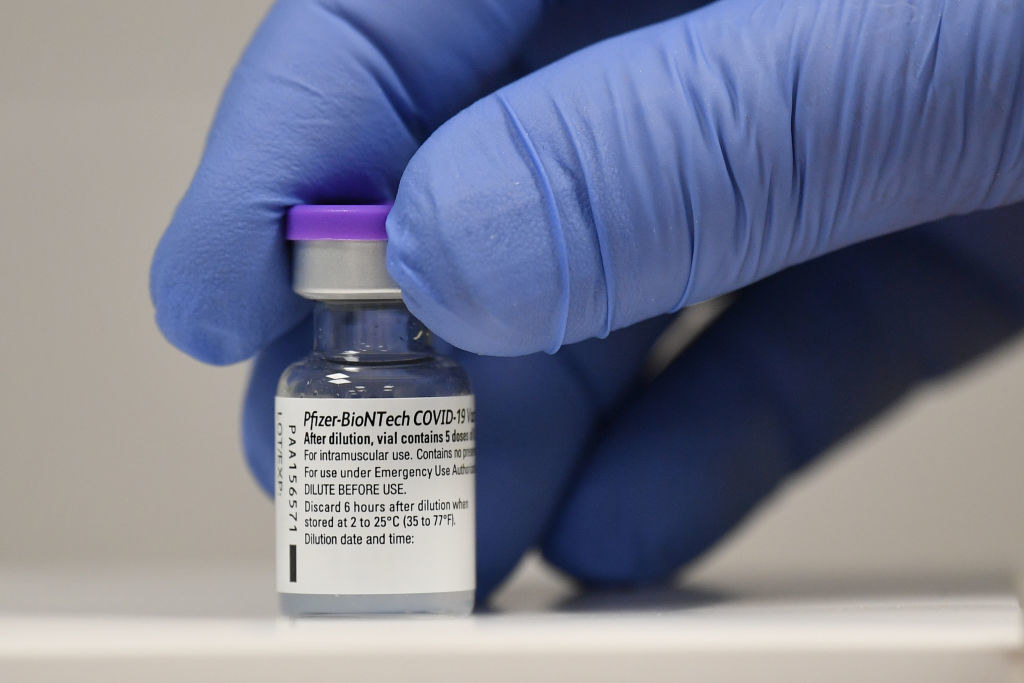 A small vial reading &quot;Pfizer-BioNTech COVID-19 vaccine&quot; is held between two fingers of a gloved hand