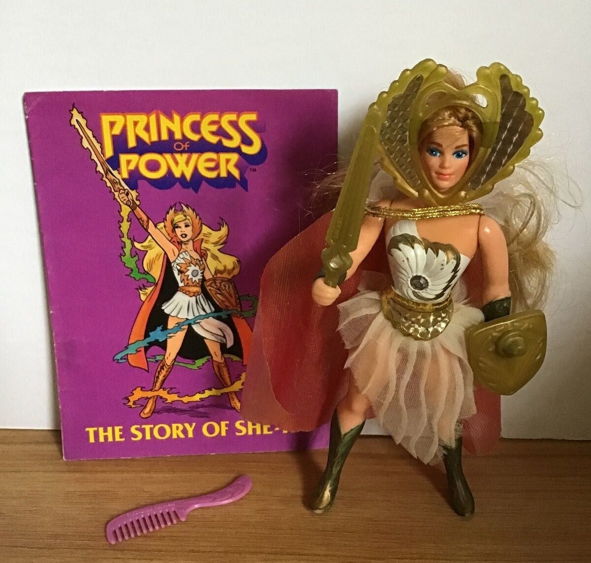 A She-Ra action figure holding her sword and shield standing next to a purple She-Ra comic  