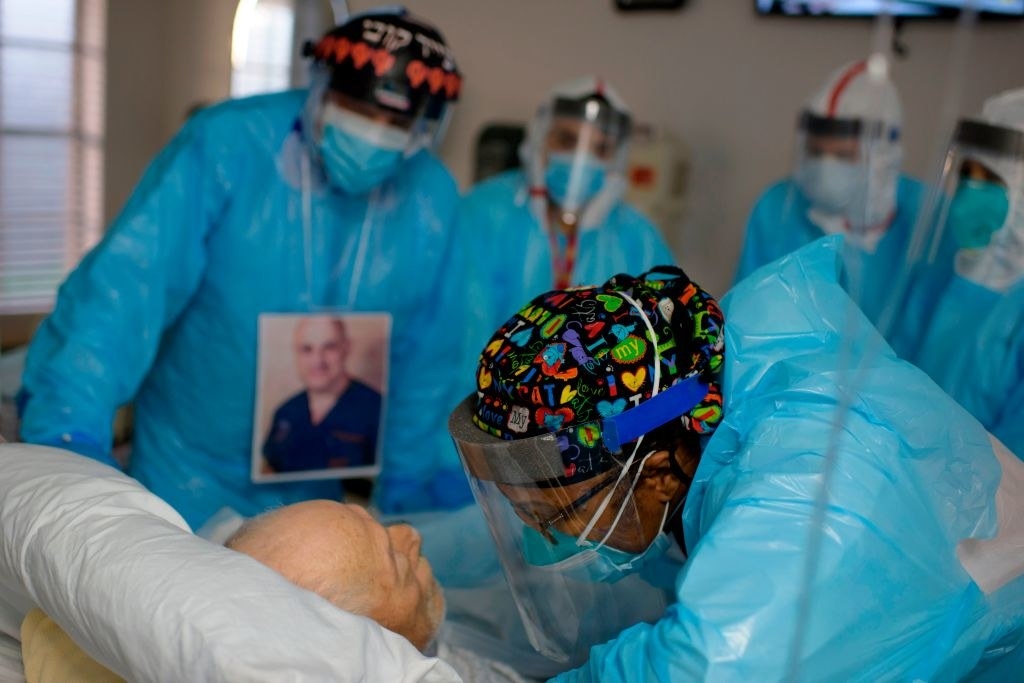 A group of healthcare workers in blue uniforms stand around a hospital bed; one woman wearing a plastic face shield leans over and talks to an older man who is lying in the bed