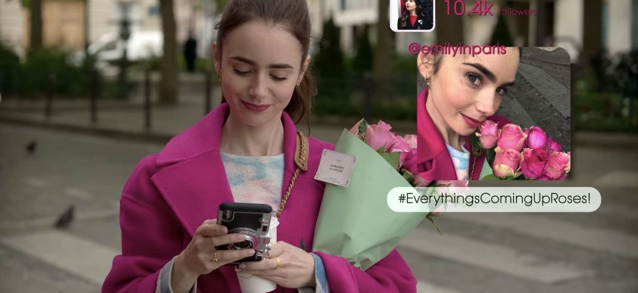 Emily looking at her phone; she has posted a picture of her with roses, captioned #EverythingsComingUpRoses!