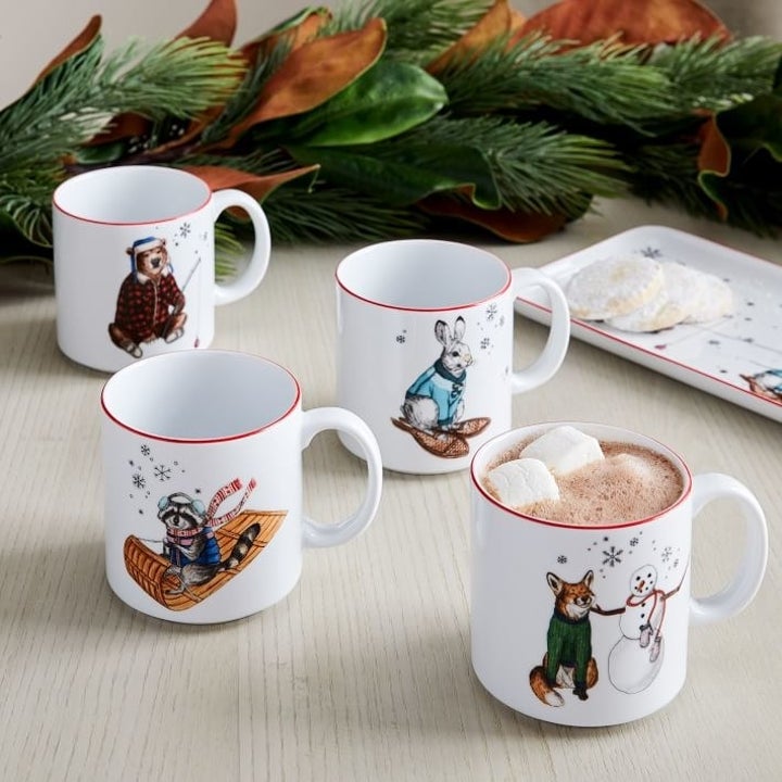 White mugs with red piping around the rim and holiday-themed images of animals sledding, building snowmen, and fishing