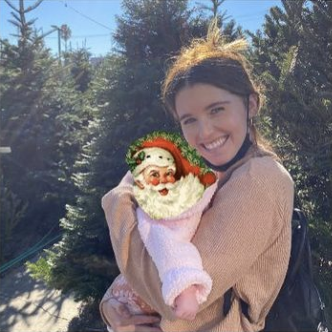 Katherine Schwarzenegger stands in front of Christmas trees, holding a baby, whose face is covered with an image of Santa Claus&#x27;s face with a hat and beard