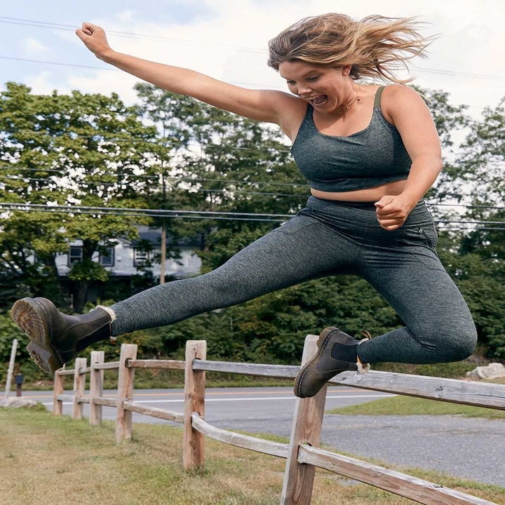A model in green heather hi-rise leggings and a matching sports bra, jumping over a fence