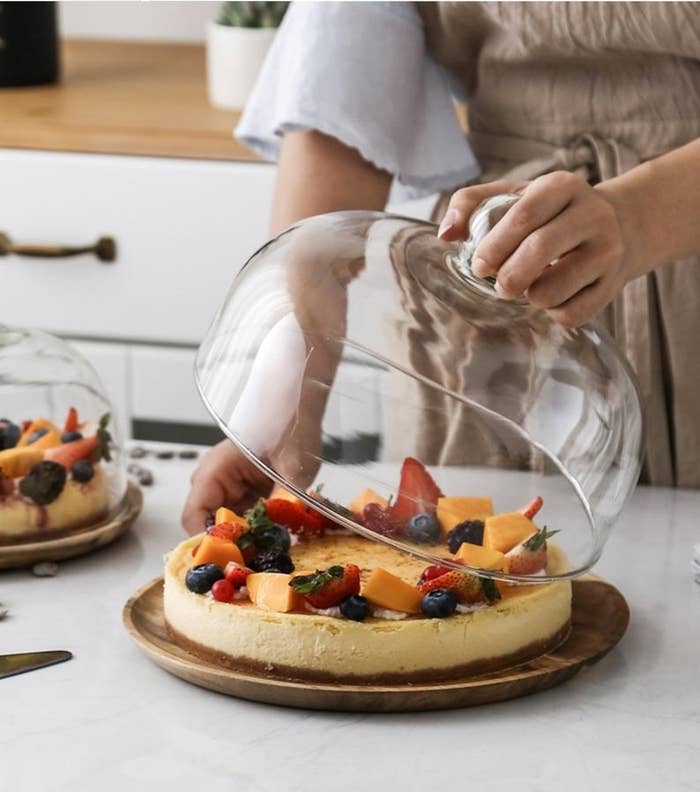 A hand lifting the glass lid off of a large walnut cake stand