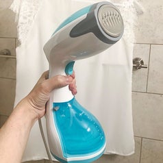 A reviewer photo of a hand holding out the blue and white handheld steamer 