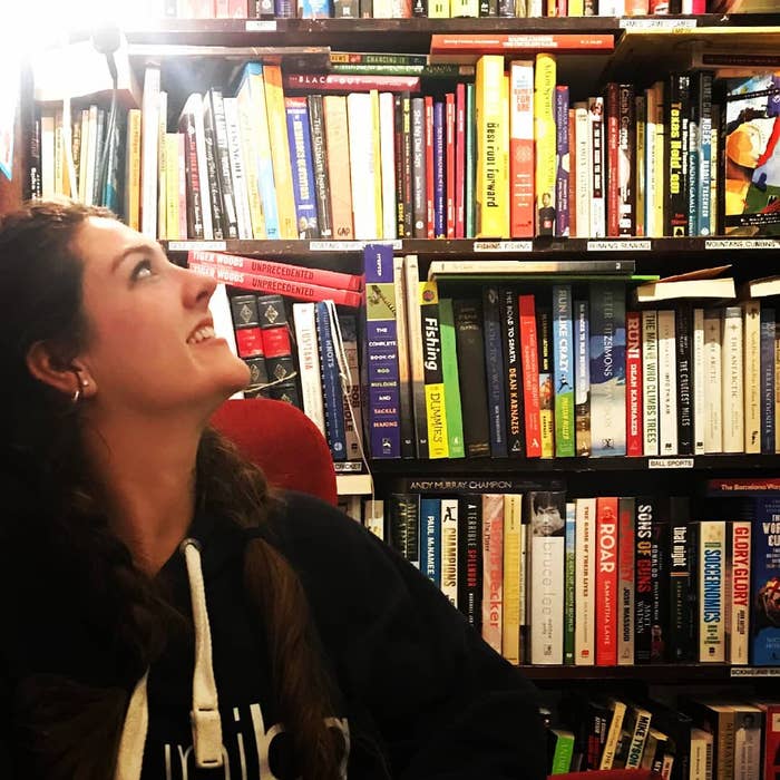 Person sits in front of a wall of books looking at the titles and smiling