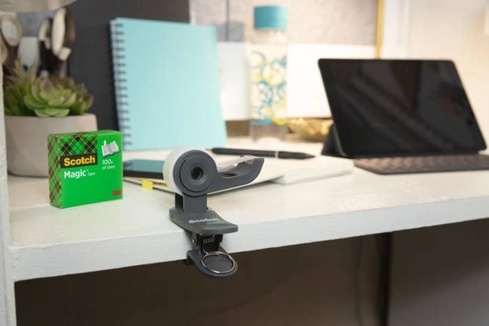 Black tape dispenser on a desk next to a green package of Scotch Magic Tape