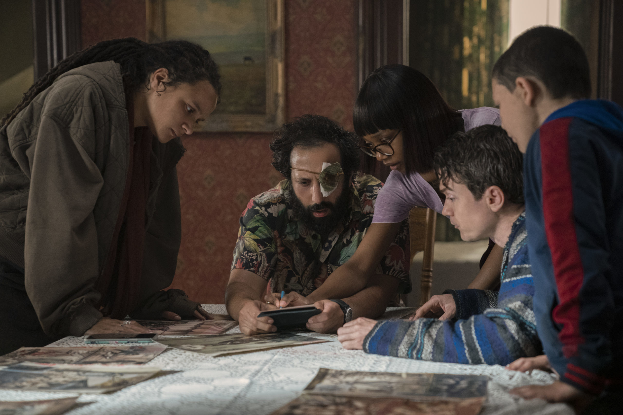 The characters from &quot;Utopia&quot; sitting around a desk that is covering in photos