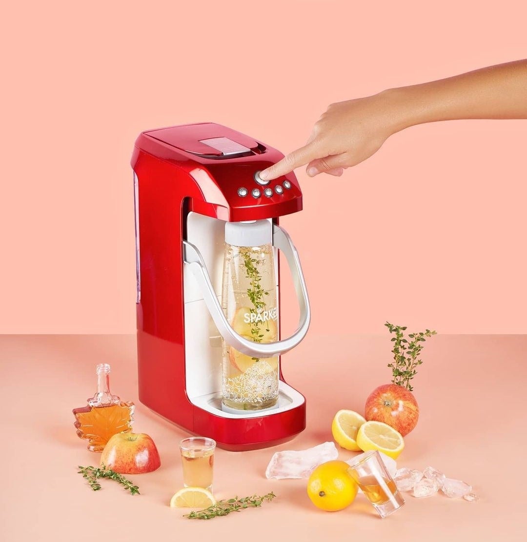 A person pressing the top button on the sparkling water maker, surrounded by apples, maple syrup, herbs, and lemons