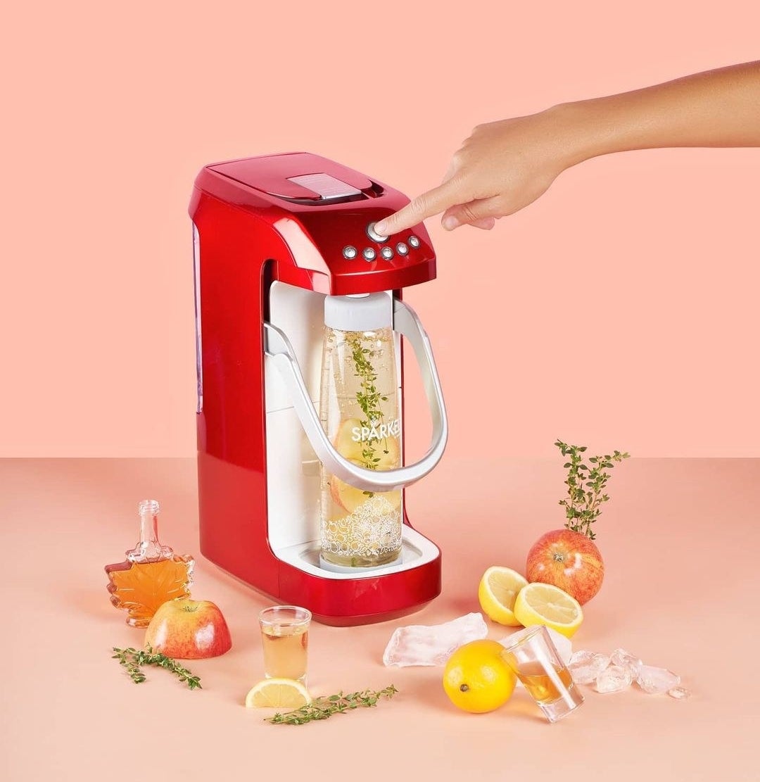 A person pressing the top button on the sparkling water maker, surrounded by apples, maple syrup, herbs, and lemons