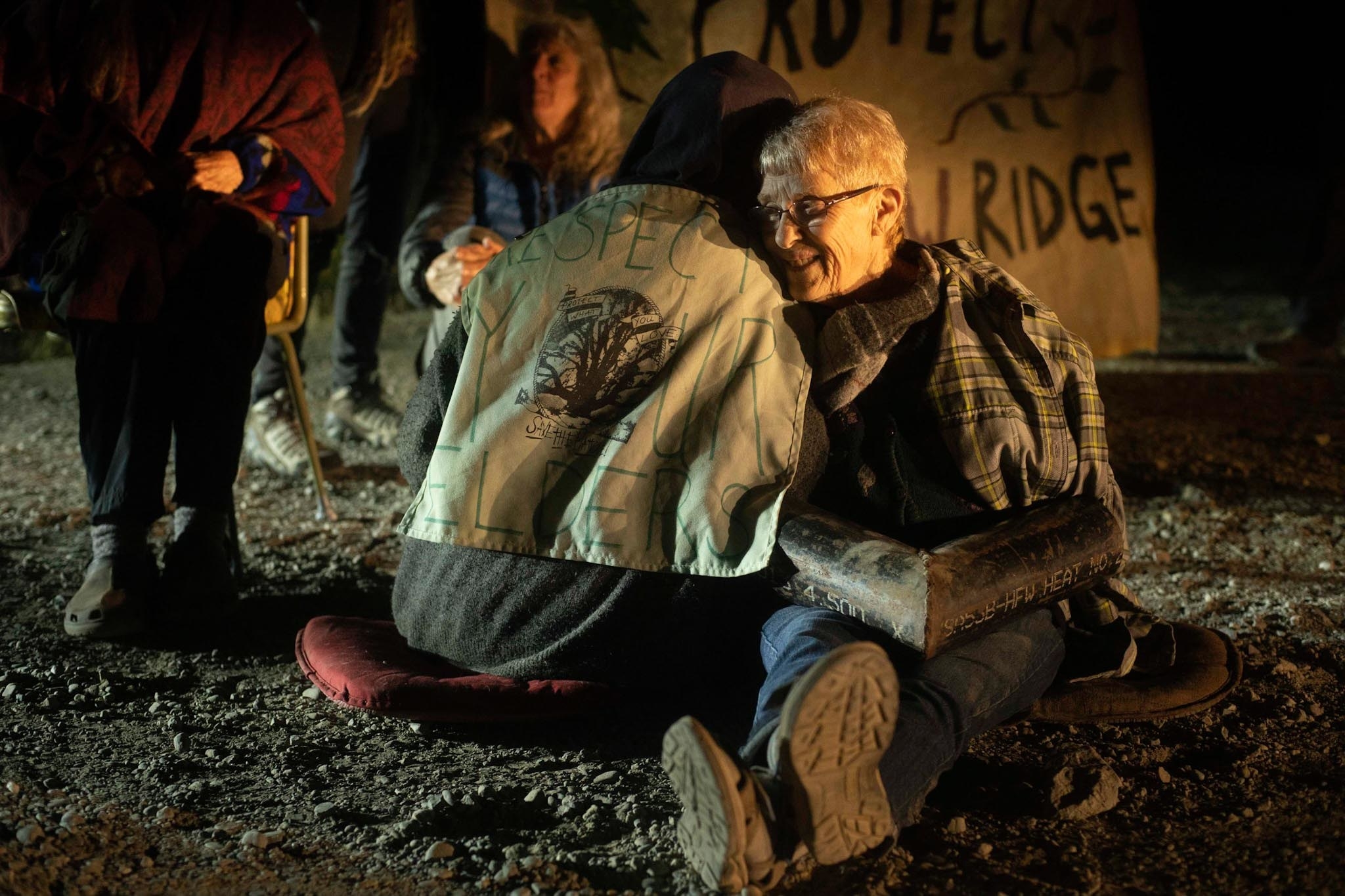 An older activist with their arms in a lock is hugged by another activist