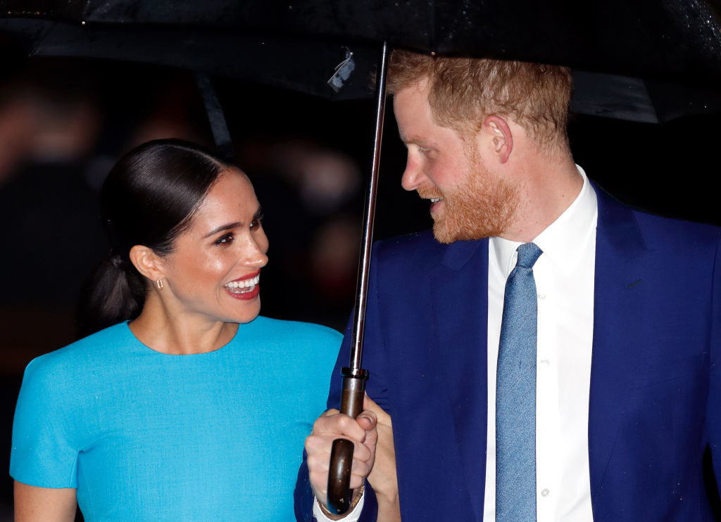 Prince Harry and Meghan Markle smiling and looking at each other under an umbrella 