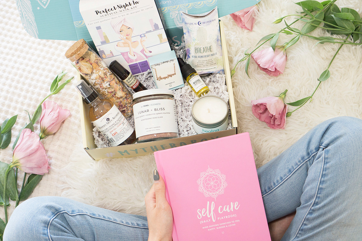 A model holding a book titled &quot;Self care&quot; siting in front of a box filled with various wellness products 