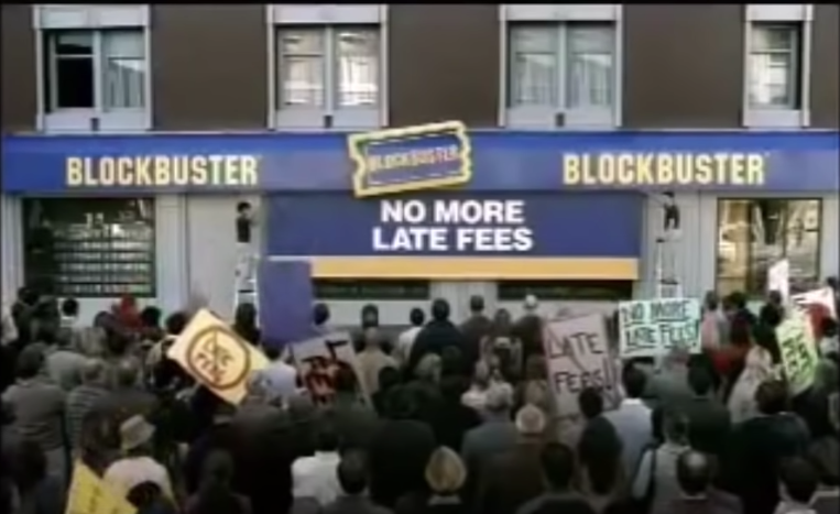 A screenshot of crowd in front of Blockbuster store with a sign that is being put up that says &quot;No More Late Fees&quot;