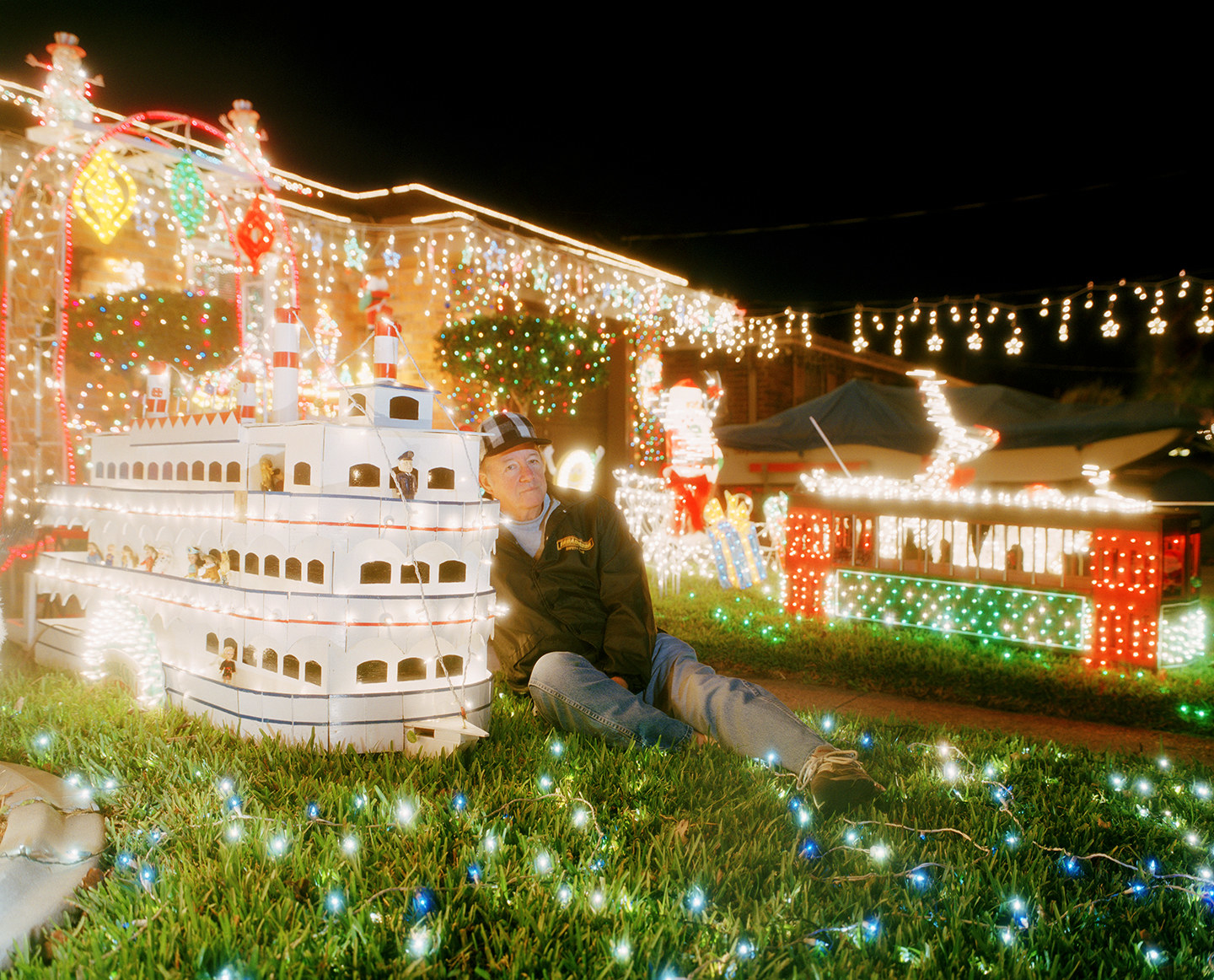 A man and a few models of riverboats, on the lawn of a house covered in Christmas lights