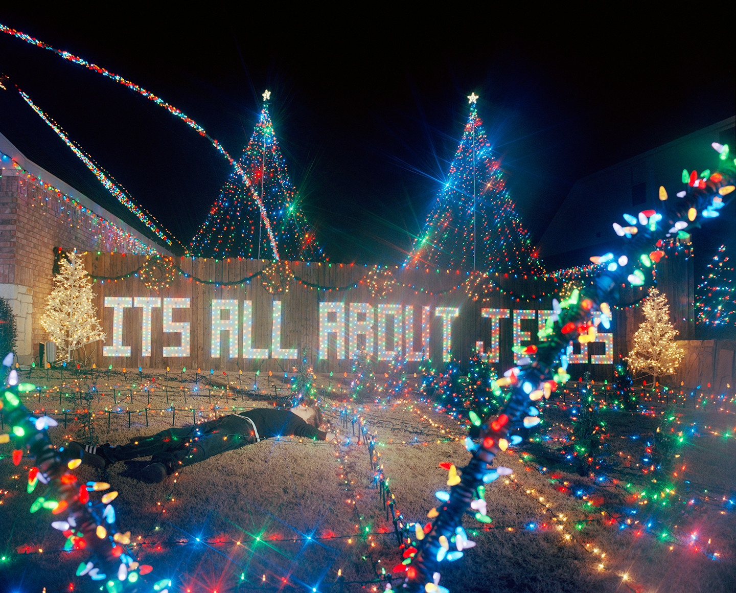 A child lying on a lawn in front of a sign and amidst Christmas lights