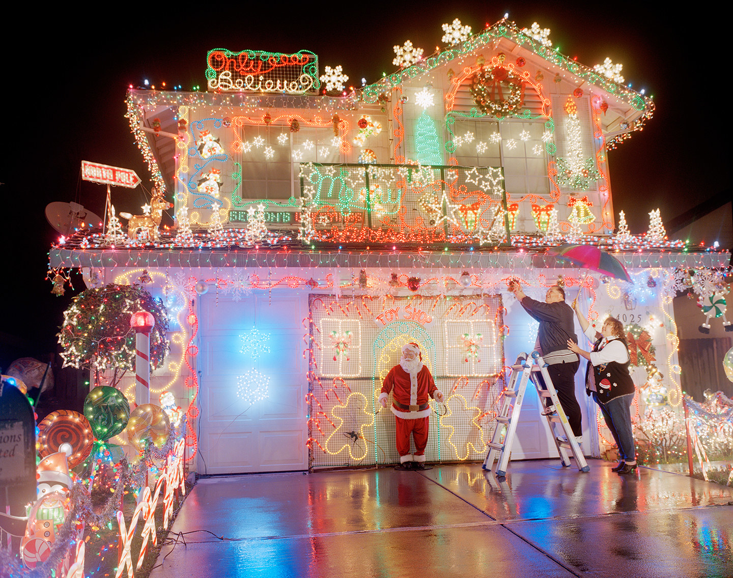 A man on a ladder adjusting Christmas lights on a house, with a woman supporting him, and a model of Santa