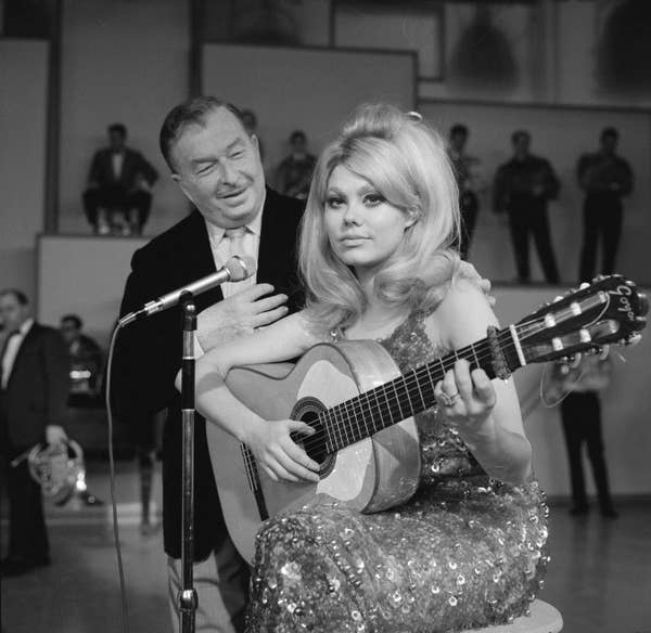 Charo on a stage sitting on a stool and in front of microphone while holding a guitar 